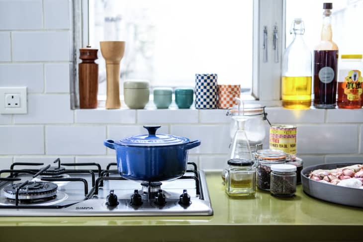 Blue pot on stove, jars of spices, and cooking oil bottles on a green kitchen counter near a window.