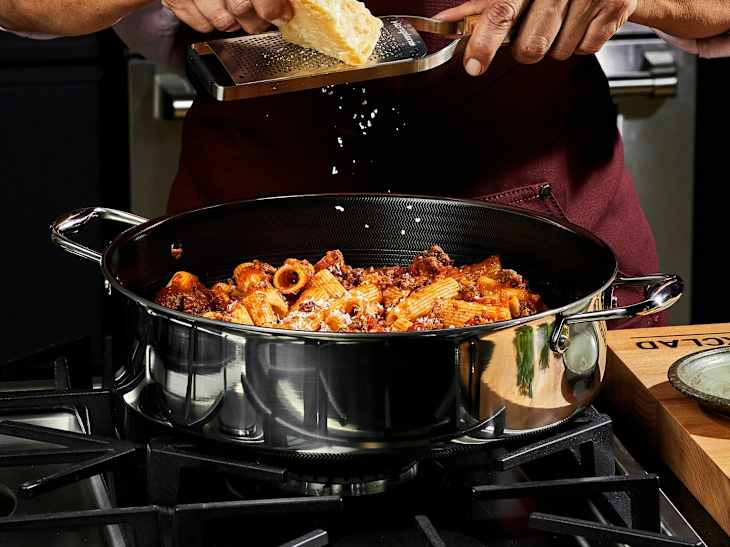 A person grating cheese over a pot of rigatoni pasta with meat sauce on a stovetop.