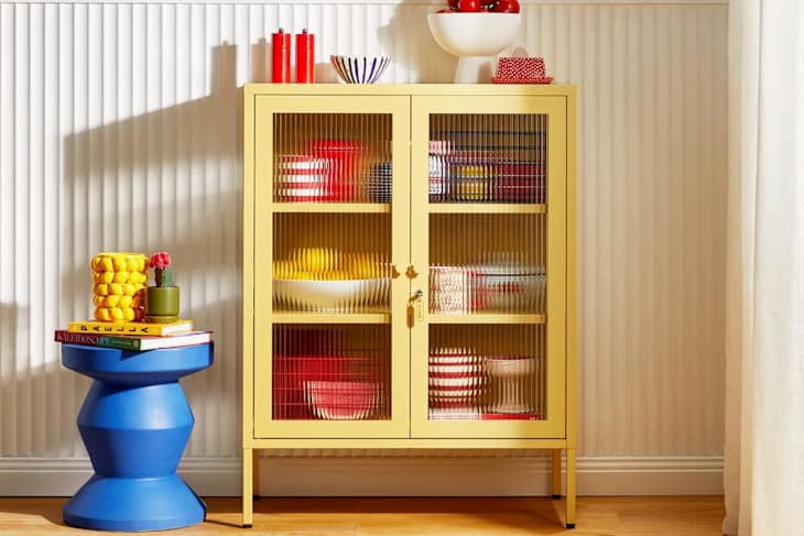 A yellow cabinet with glass doors displaying colorful dishes, next to a blue side table with books and a plant.